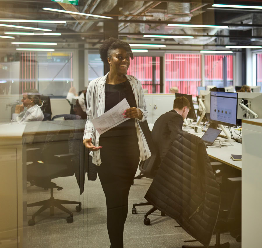 A woman wearing a blazer walking through an office carrying papers whilst smiling. People are working in the background on their computers.