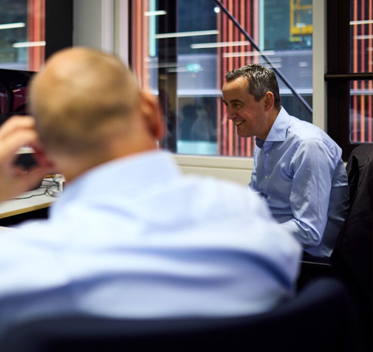 Two men in a lit up office smiling at each other. They are next to a window, creating a professional and relaxed work setting.