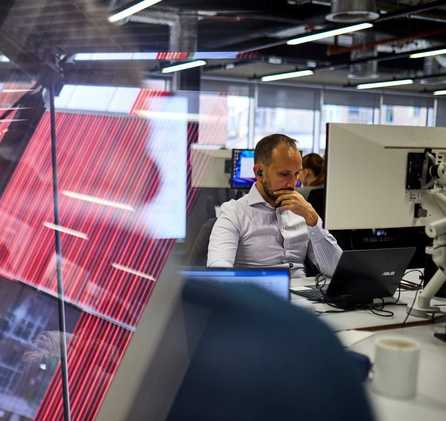 A man wearing headphones, focusing on his computer, with reflective red stripes on the glass. A modern workspace in a tech environment.