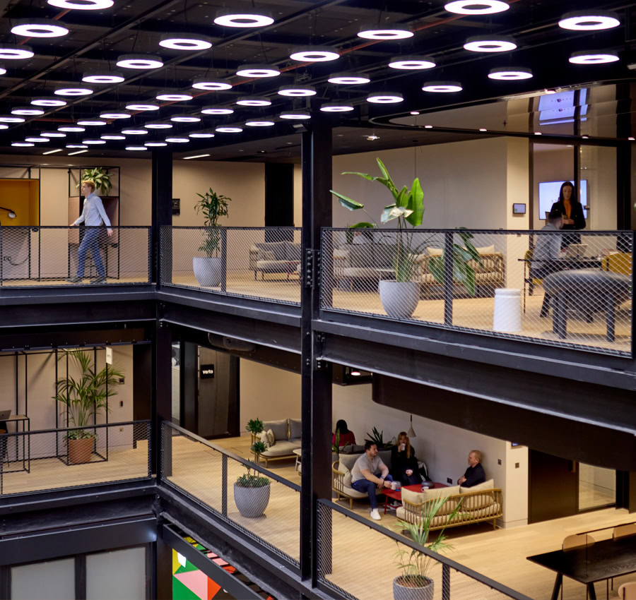 A modern office space with open balconies, and ceiling lights. THere are plants and people seated on different levels.