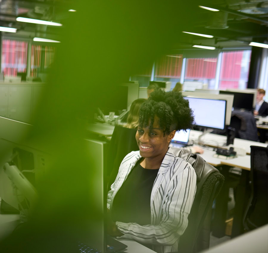 A smiling woman in a striped blazer sitting in an open-plan office, obscured by greenery in the foreground.