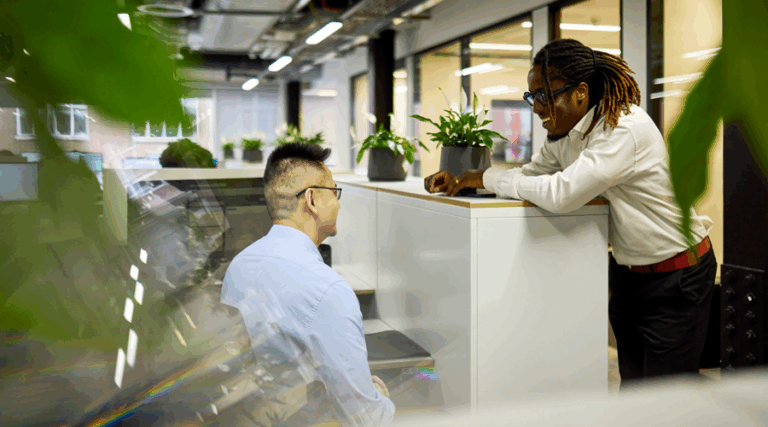 A modern white office with windows and light with green plants on desks. Two workers talking in suits and looking at a computer.