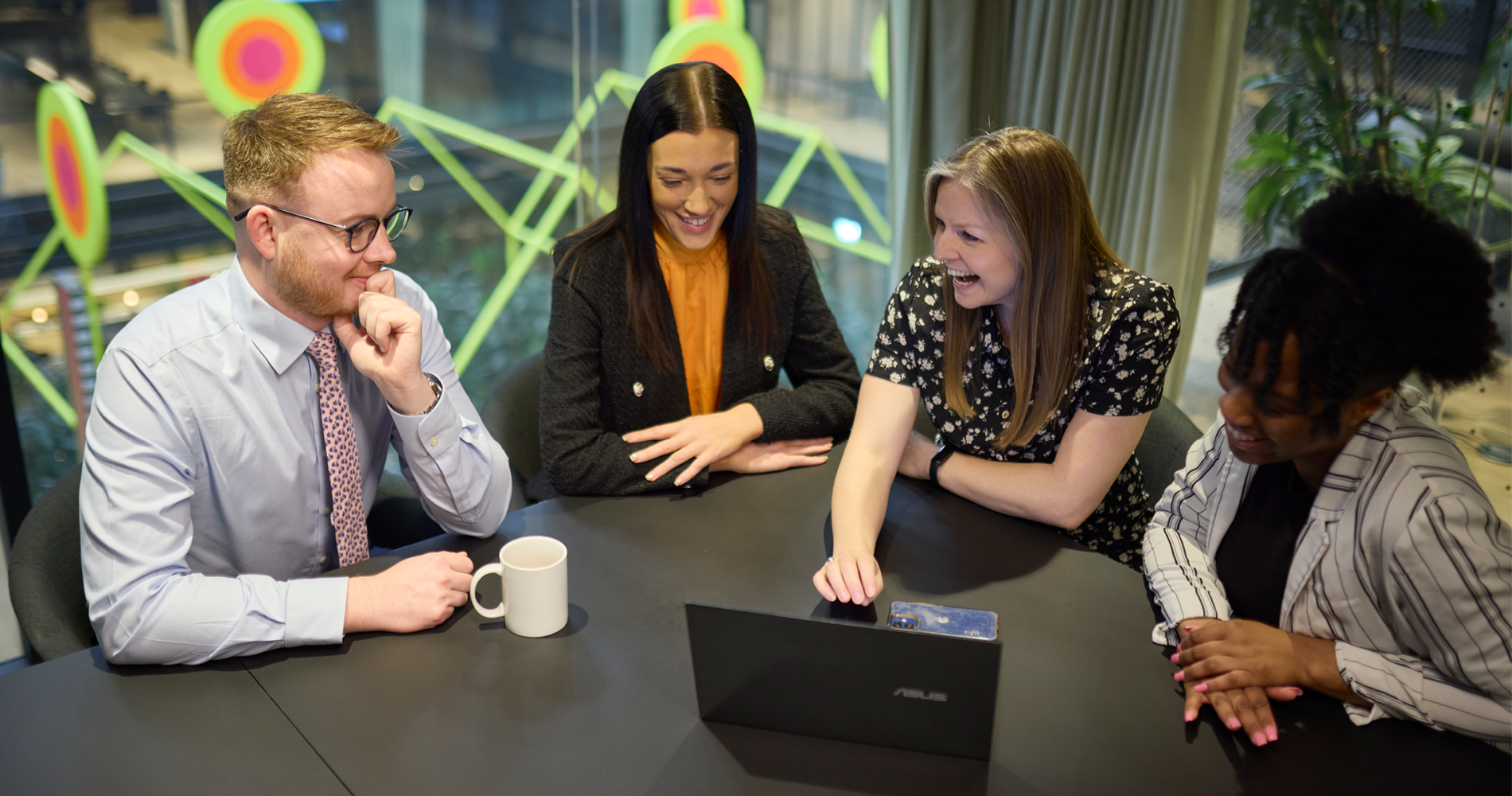 A group of workers in a meeting hub looking at a computer, surrounded by windows and plants. Featuring vibrant and colourful art in the background, creating an energetic and modernised finish.