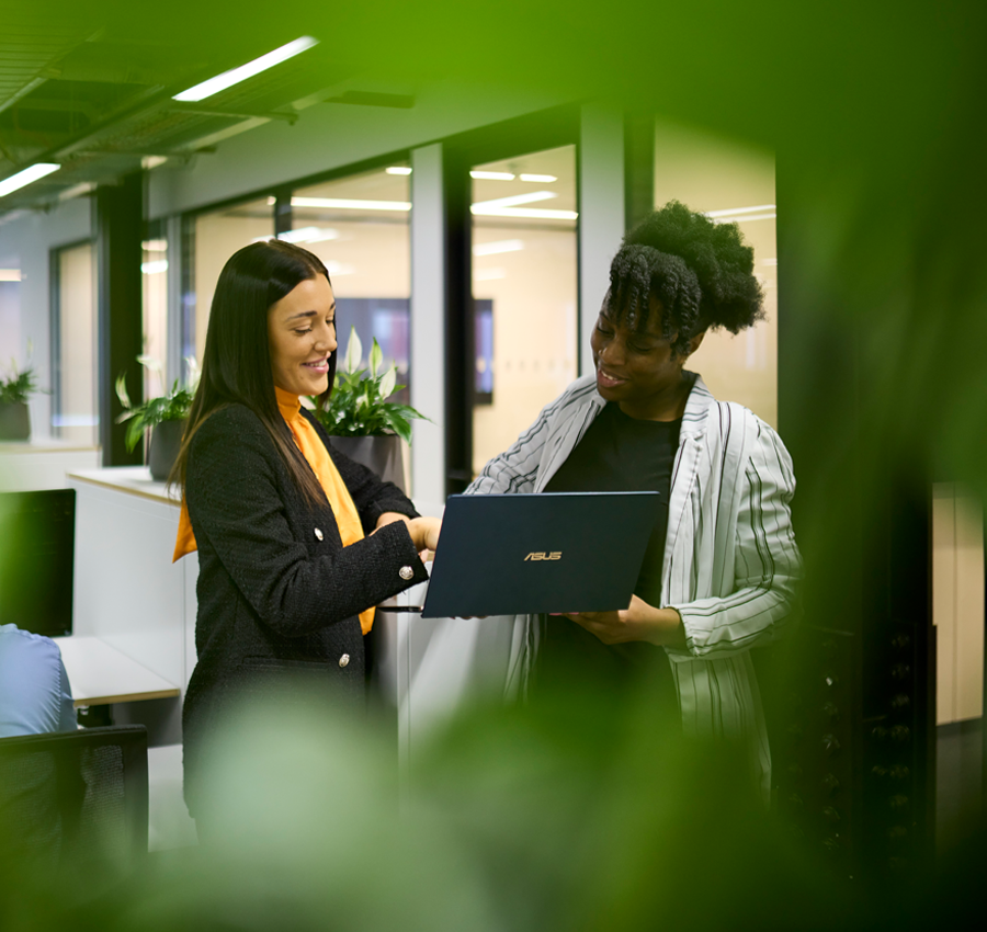 Two workers looking at a computer in a white modern office with green plants and computers.