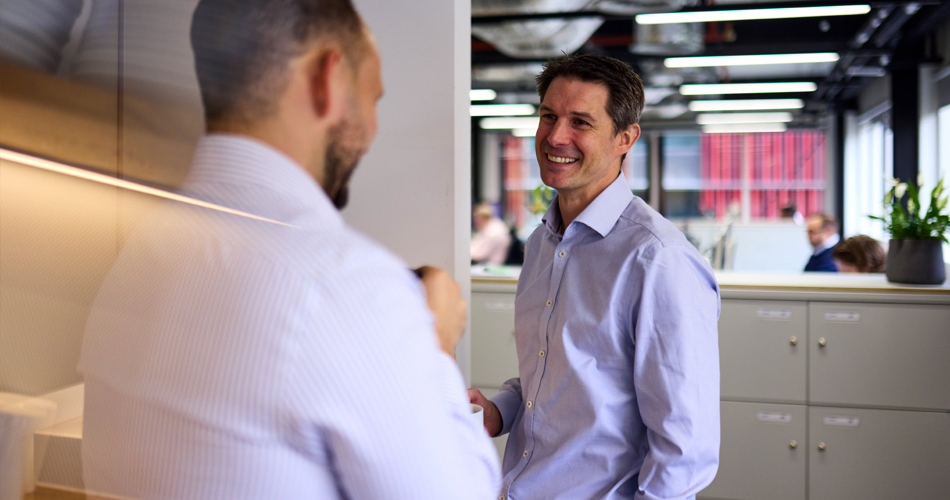 Two men in an office having a conversation. The man facing the camera is smiling, creating a professional environment.