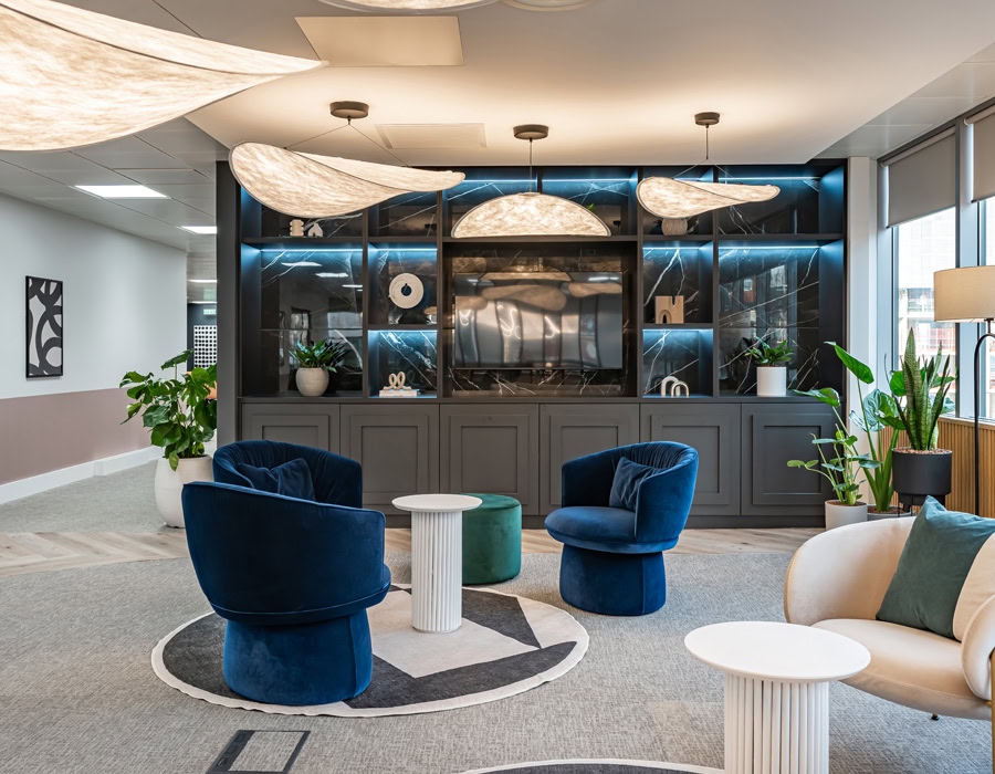 Modern lounge with blue and beige chairs, white round table, geometric rug and wall shelving. Soft lighting creates a cosy atmosphere.