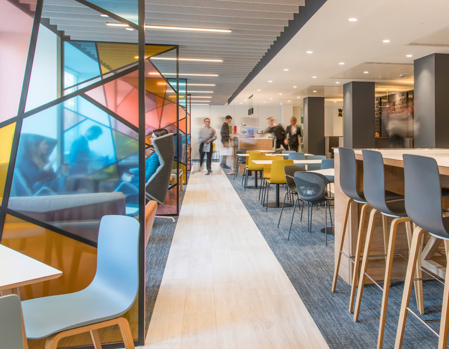 A modern office lounge with colourful geometric glass partitions, wooden accents and varied seating. People walking and chatting in the background.