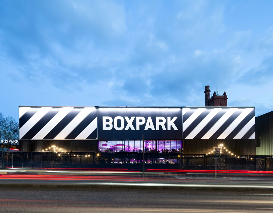 BoxPark sign in bold and capital letters with stripes on a building at dusk. Red light trails from passing cars which adds energy to the scene.