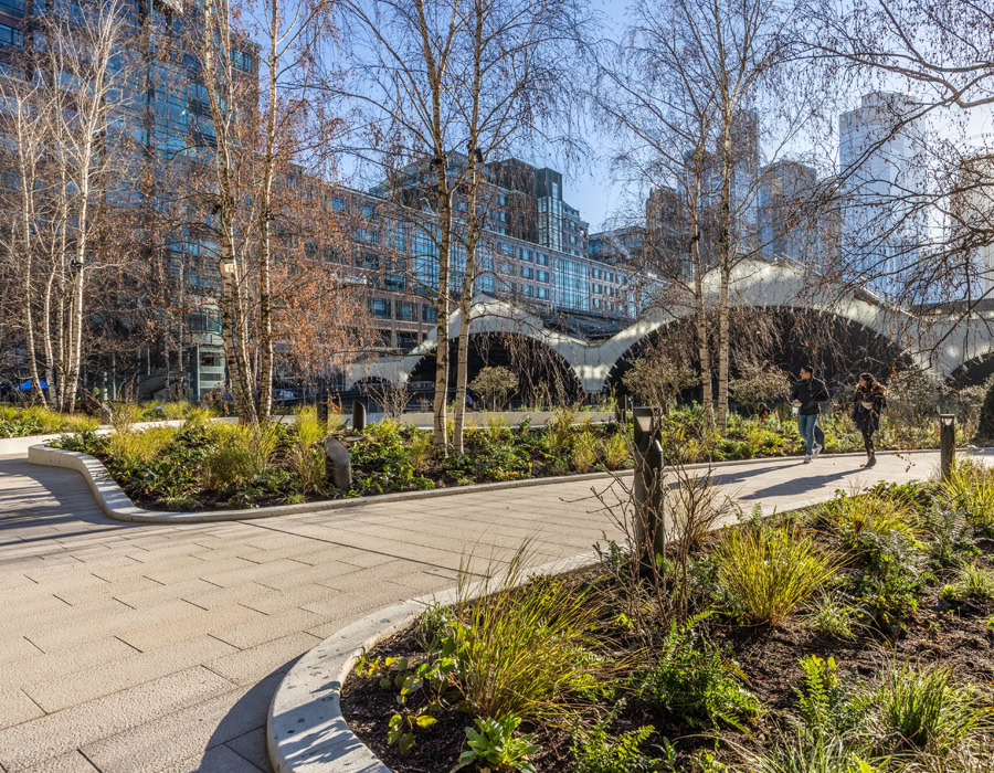 An urban park scene with a curved path, surrounded by tall trees and plants. People walk under with modern buildings in the background.