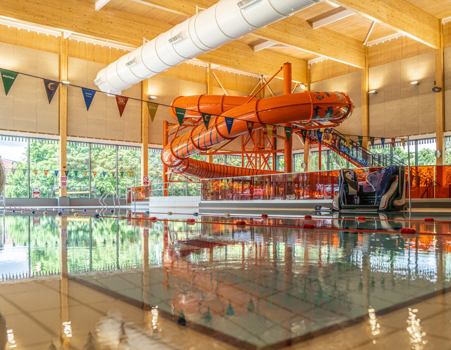 An indoor pool with an orange slide, colourful bunting and a reflective water surface. Natural light filters through large window panes, creating a bustling atmosphere.