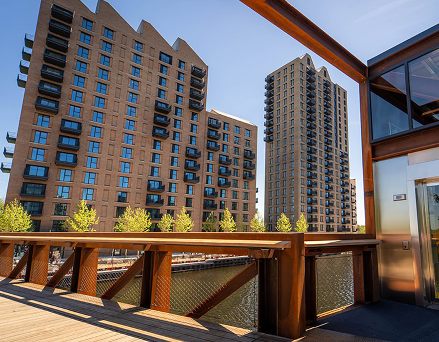 Three tall, modern brick buildings with balcony stands by a canal under a blue sky. A wooden platform with glass and metal accents overlooking the water.