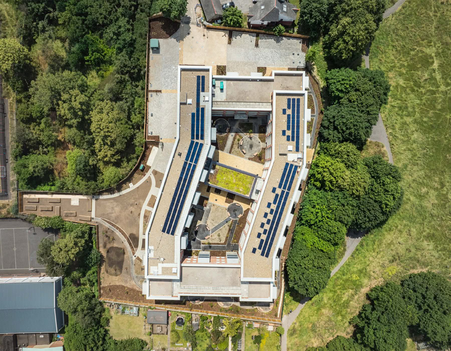 Aerial view of a large U-shaped building with solar panels on the roof, located among dense green trees and a grassy area, highlighting sustainability elements.