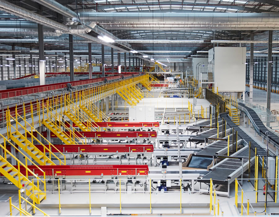A large factory interior, with conveyor systems featuring vibrant red and yellow structures, surrounded by lighting and metal structures