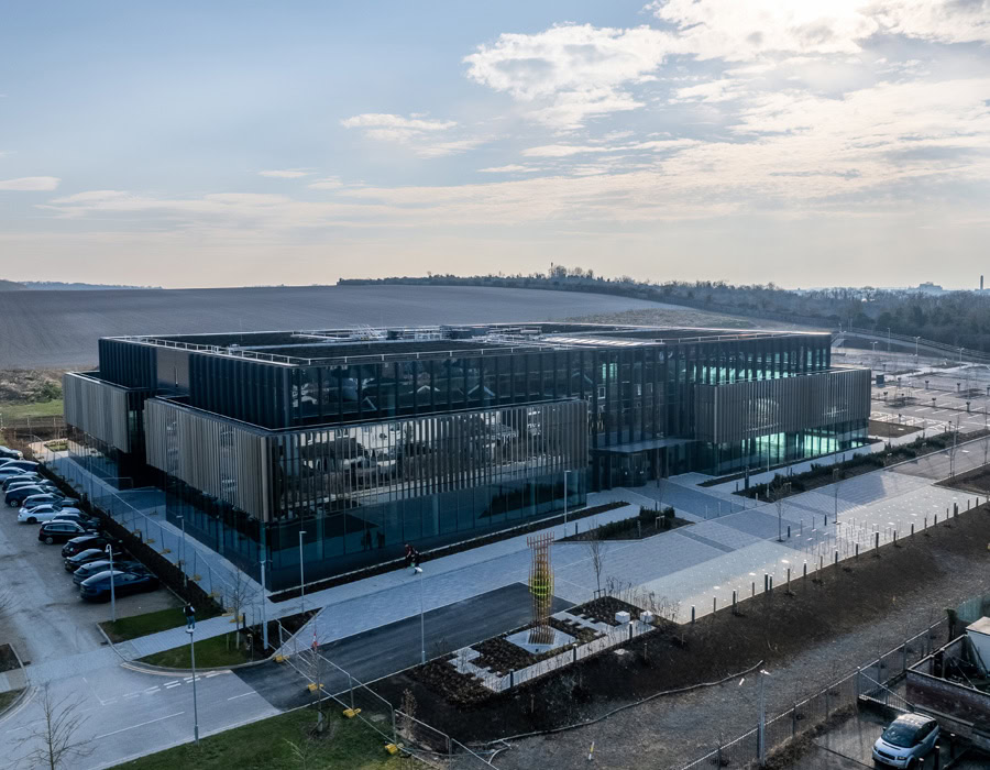 A modern, low-rise building with a glass facade and vertical metal accents in a spacious, rural landscape under a fairly cloudy sky.