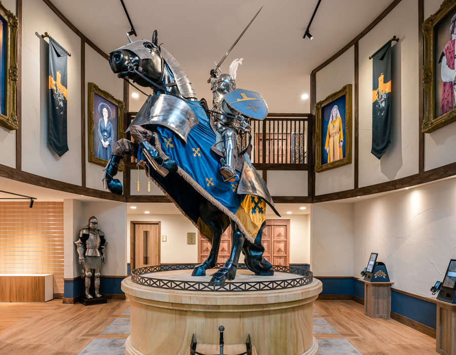 A knight in full armour on a horse statue, with a blue and gold banner, surrounded by portraits in a medieval hall.