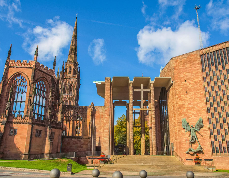 An old historic red-brick cathedral with modern glass, a tall spire and sculpture. The blue sky portrays tranquillity.