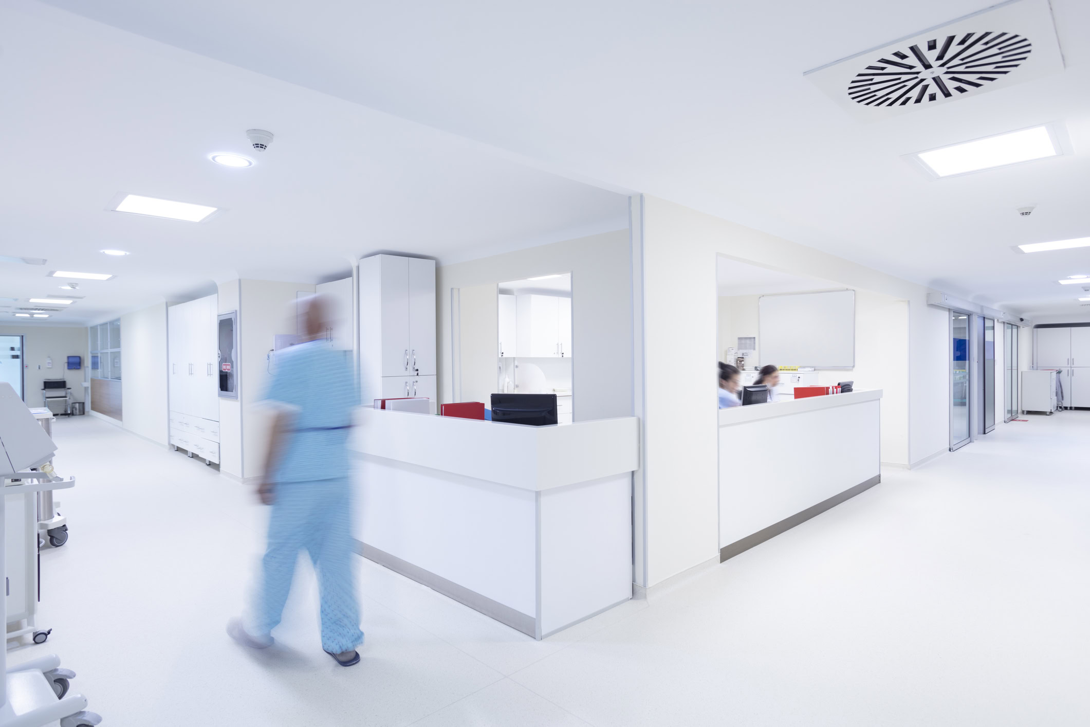 A white and clean hospital with a worker in blue walking down the isle. A reception with two nurses.