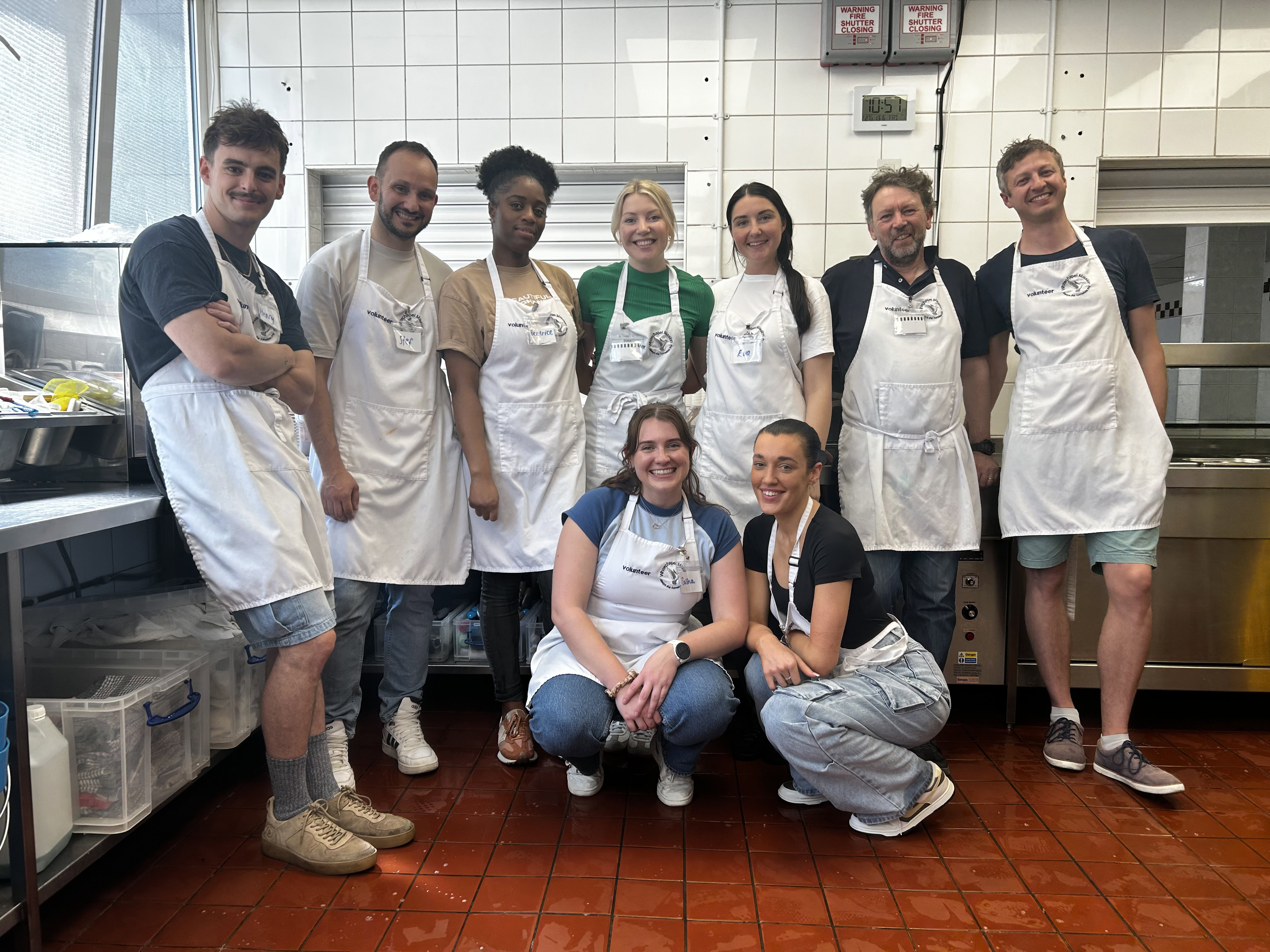 A group of people in the kitchen, dressed in aprons who are all smiling. Standing on a red tile floor portraying a collaborative atmosphere.