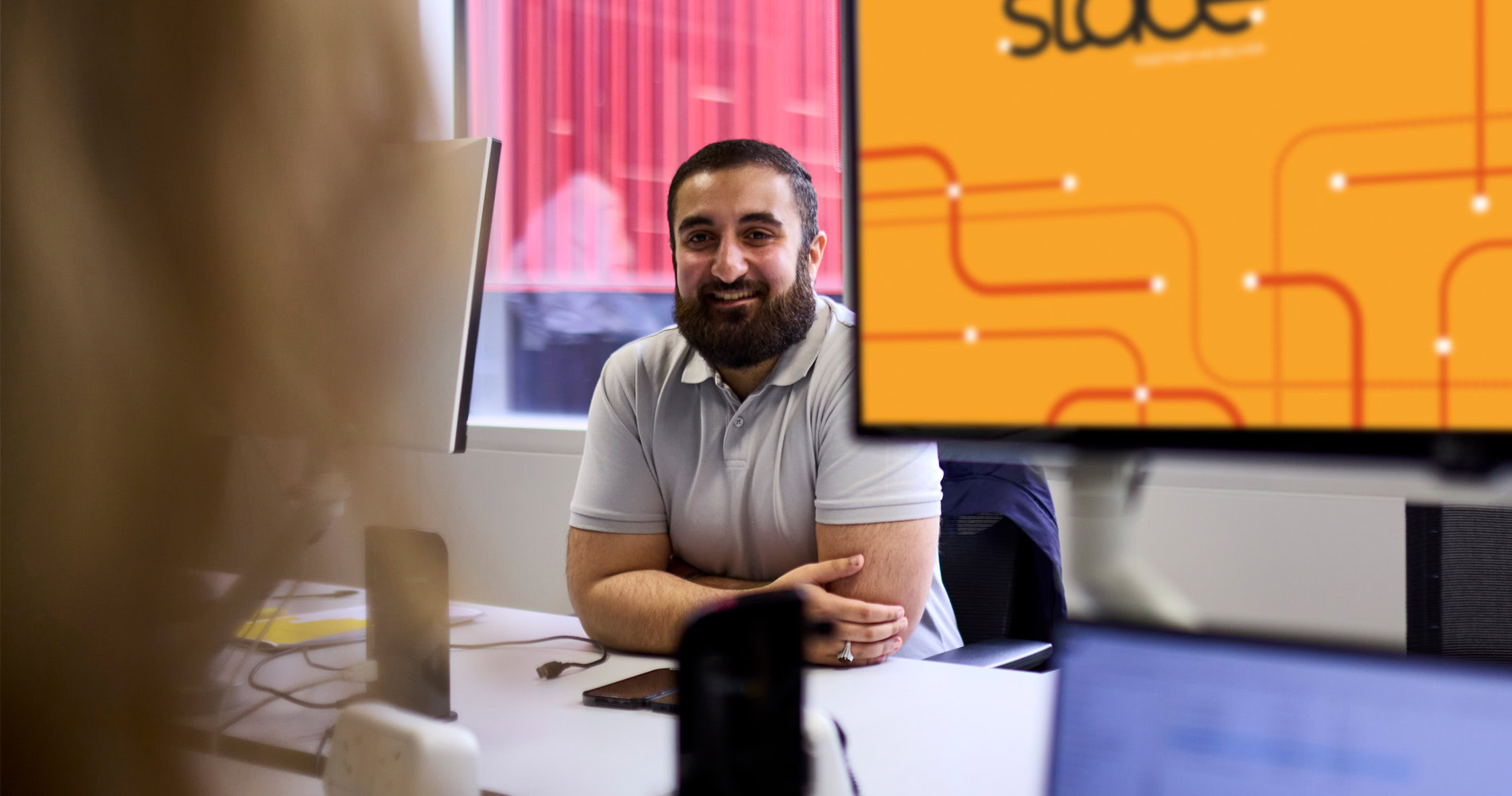 A man smiling sitting down in an office, with a computer screen showing an orange graphic in the foreground. The environment appears to be professional.