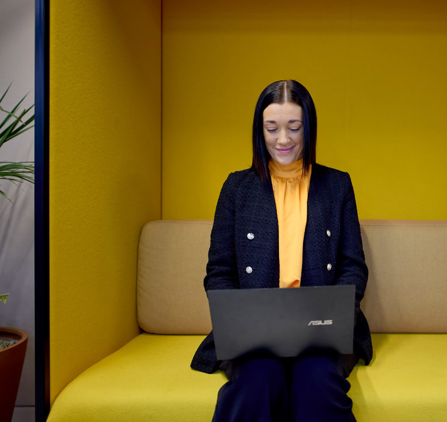 A lady sitting on a yellow sofa, working on a laptop. She is wearing a dark blue blazer smiling creating a positive mood.