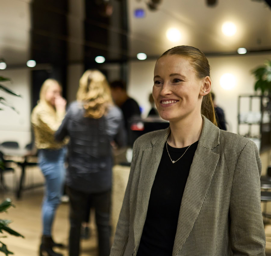 A lady smiling in a checkered blazer in a lit up office, with colleagues chatting in the background, creating a vibrant ambience