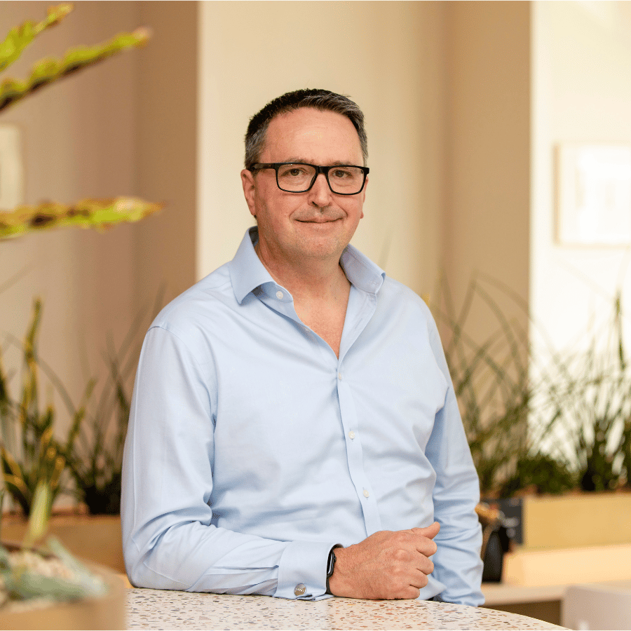 Man in glasses wearing a light blue shirt, standing next to a countertop in a modern indoor setting with plants in the background.