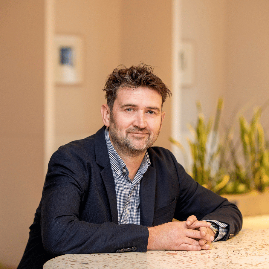 Man wearing a dark blazer sitting next to a countertop in an indoor setting.