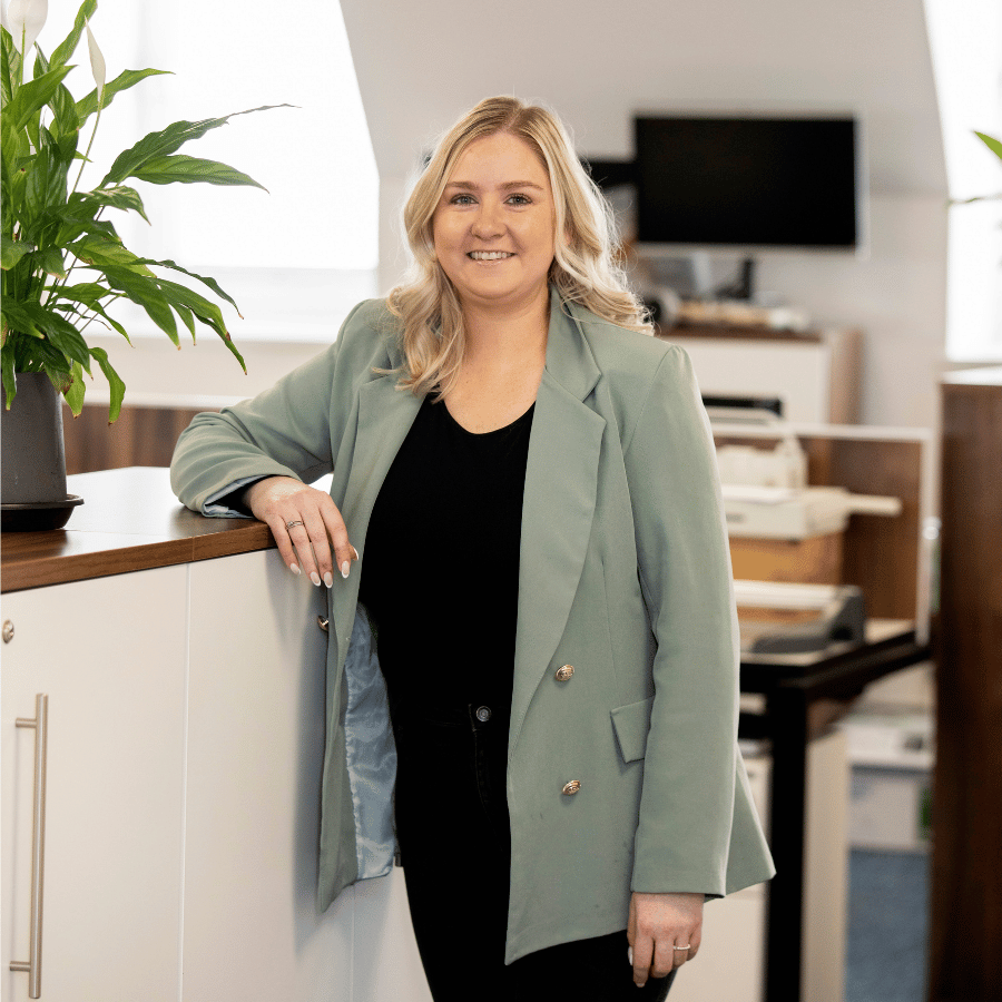 Lady smiling wearing a light green blazer and black outfit, leaning on a wooden counter in an office space.