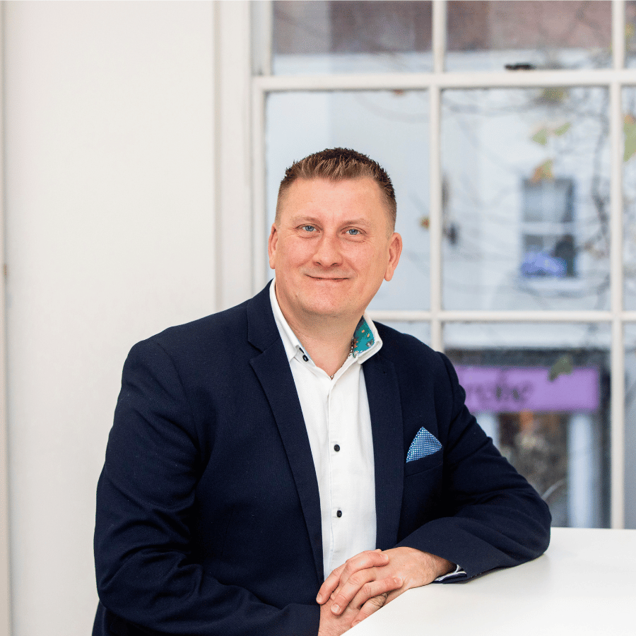 Man wearing a dark blazer and white shirt with a patterned pocket square, seated indoors near a large window.