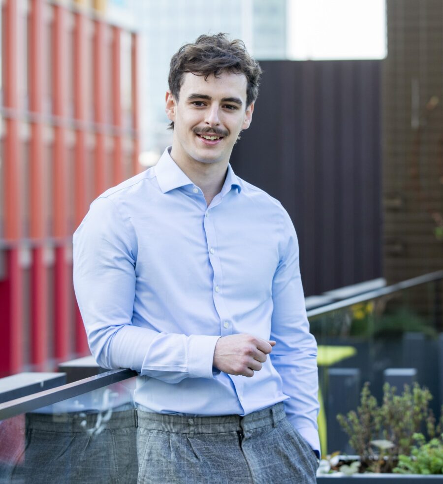 Man in blue shirt and grey trousers standing against rooftop railing with office buildings in the background