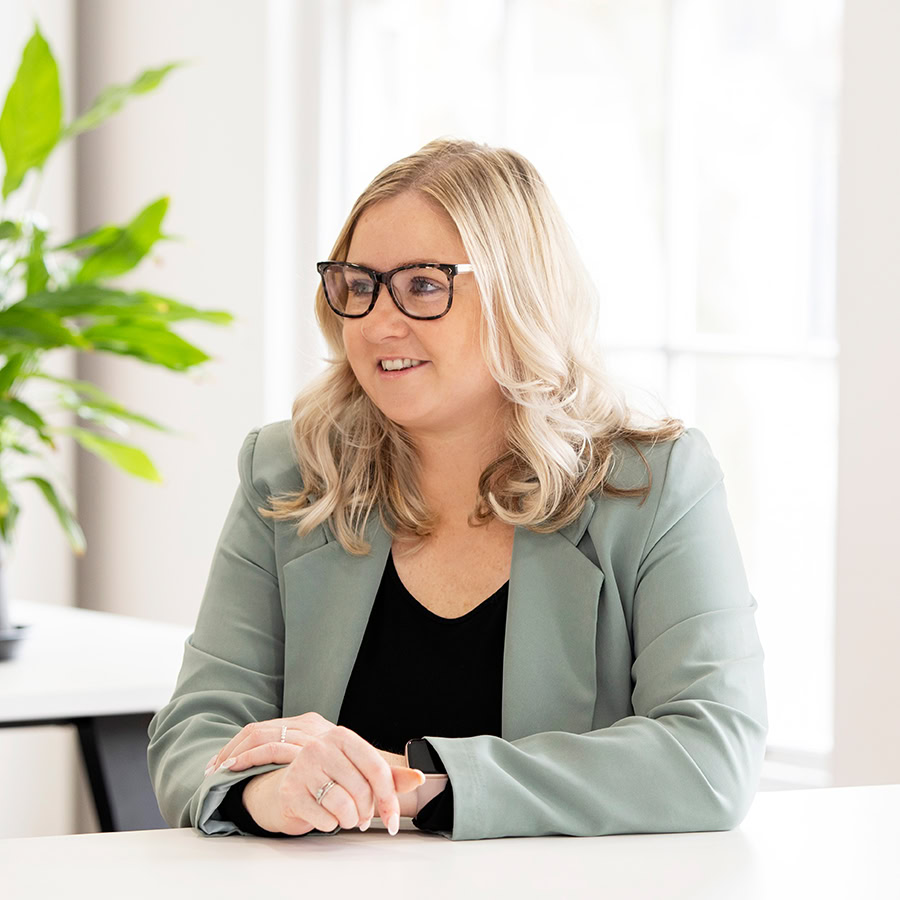 Lady wearing a light green blazer over a black top, standing in a modern office setting near a wooden counter with a potted plant.
