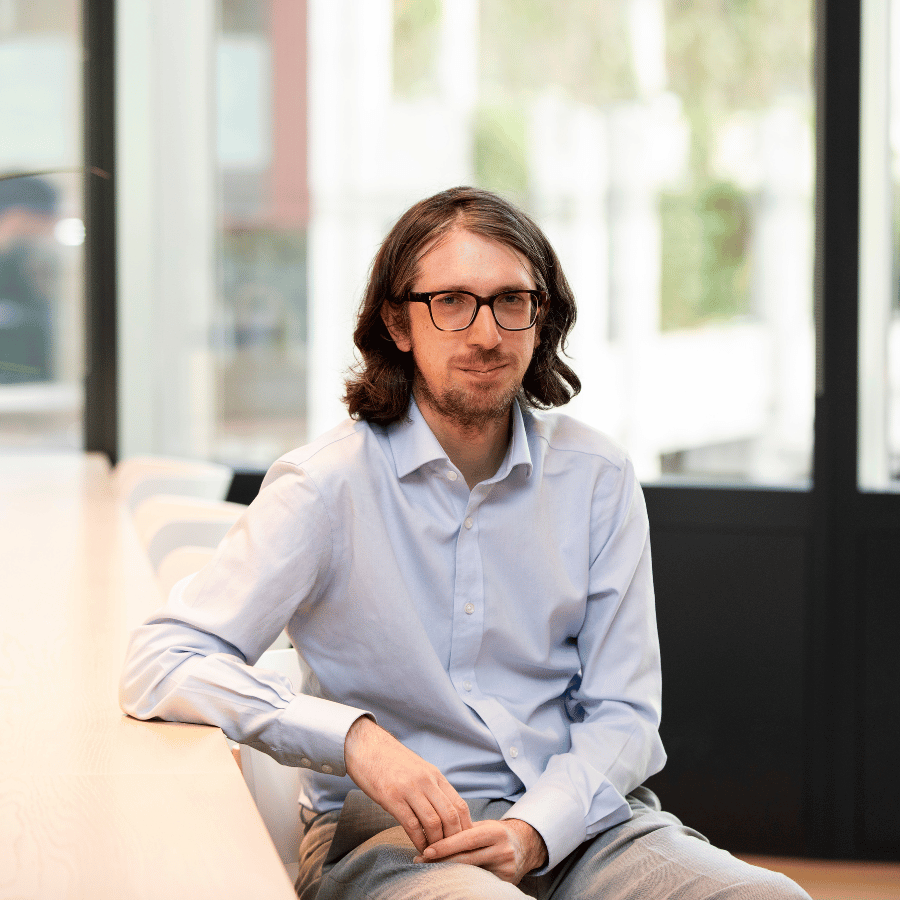 Man in glasses wearing a blue shirt, leaning his arm on a table in a modern indoor setting with windows behind.