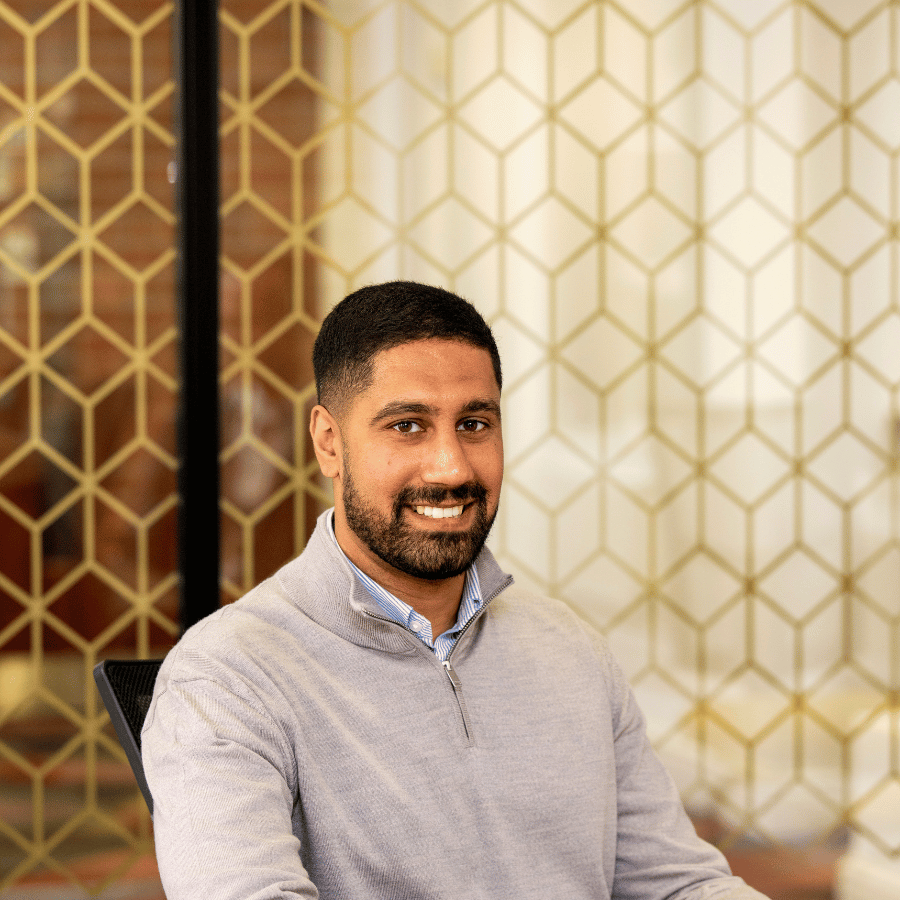 Man wearing a light grey sweater over a collared shirt, with a geometric patterned wall in the background.