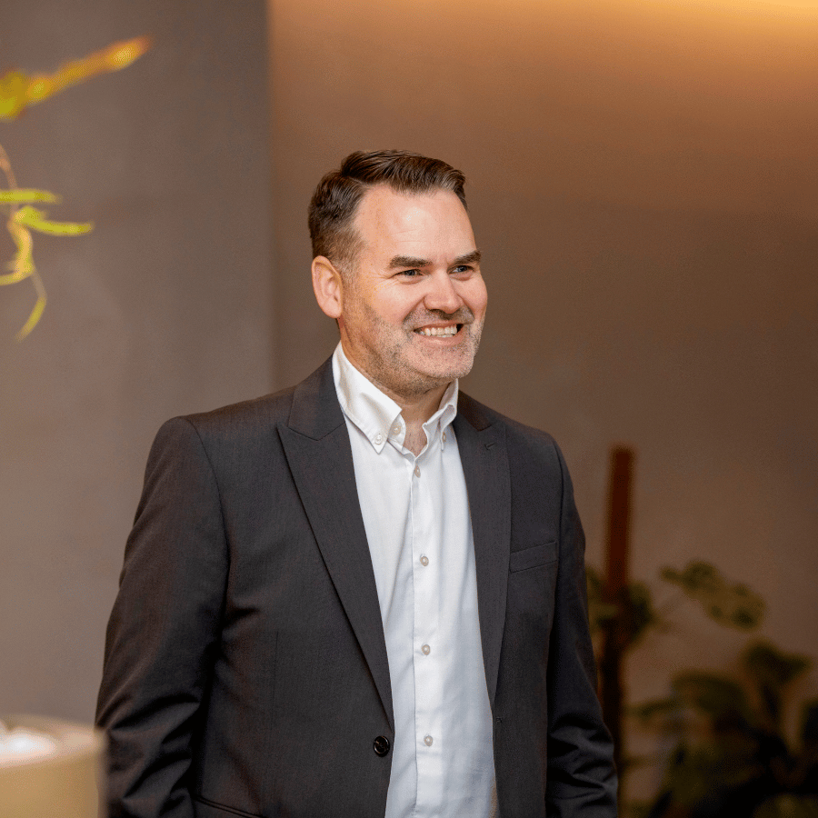 Man in a dark suit and white shirt standing indoors near plants, smiling.