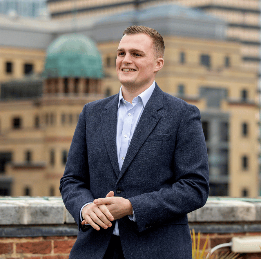 Man in a dark suit jacket and light blue shirt standing outdoors with blurred city buildings in the background.