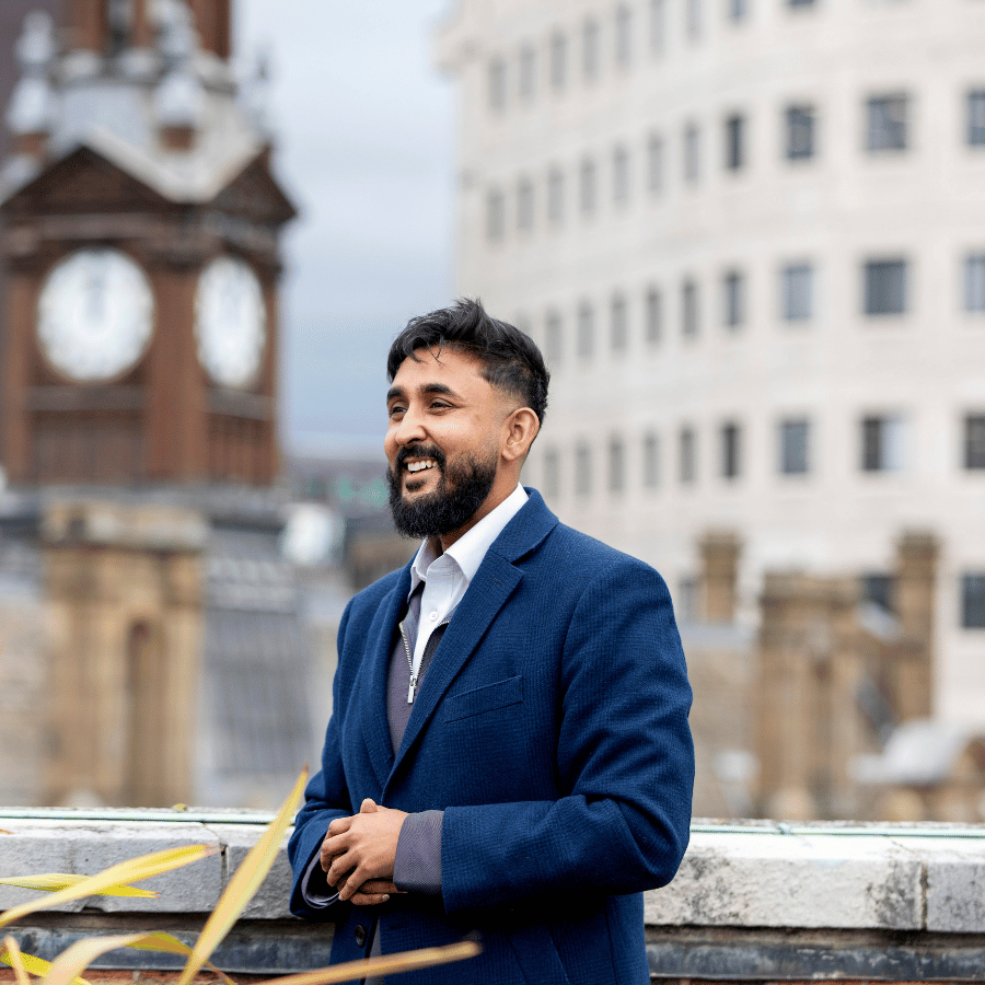 Man in a dark blue suit standing outdoors with historic clock tower and modern building in Leeds.
