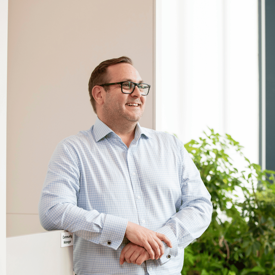 Man wearing a light blue checkered shirt, standing indoors near a white wall with a green plant in the background.
