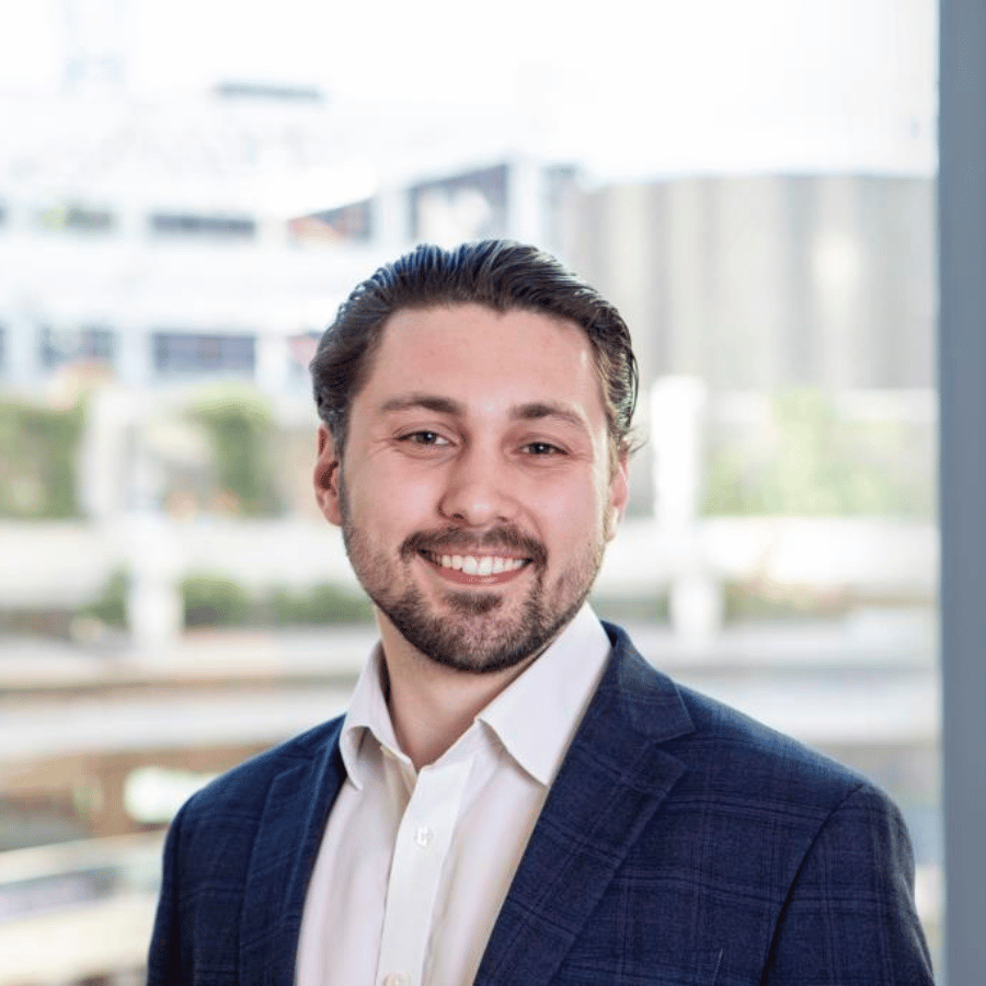 Man wearing a dark checked blazer and light-colored shirt standing indoors with large windows and an outdoor view in the background.