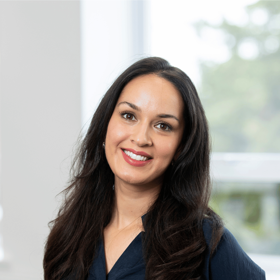 Lady with long dark hair wearing a dark top, standing indoors near a bright window with greenery visible outside.