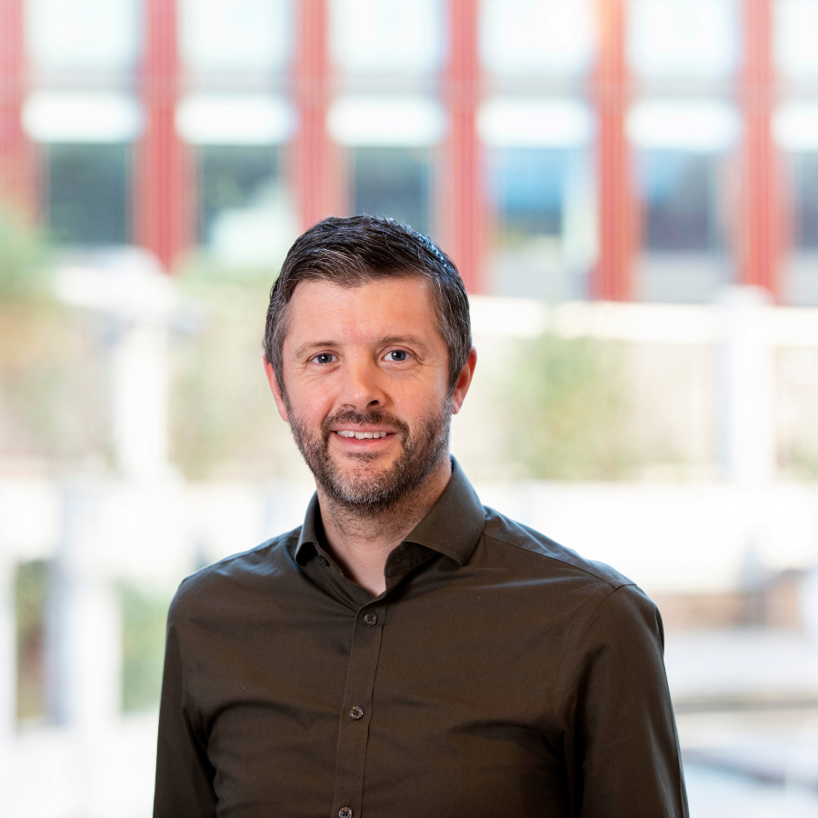 Man wearing a dark button-up shirt standing indoors with large windows and a blurred outdoor view in the background.