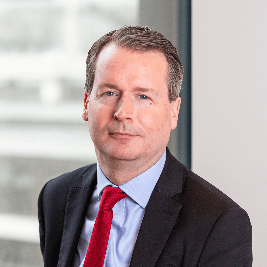 Man in a dark suit with a light blue shirt and red tie, seated indoors near a window.