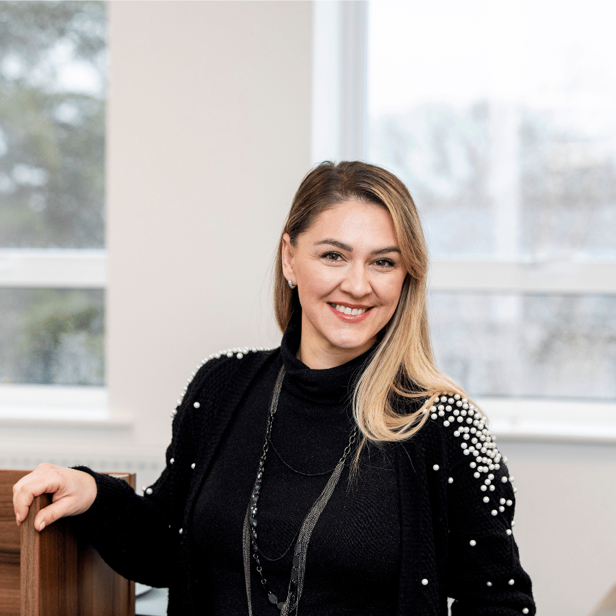 Woman wearing a black outfit with pearl details, standing indoors near a wooden surface and large windows.