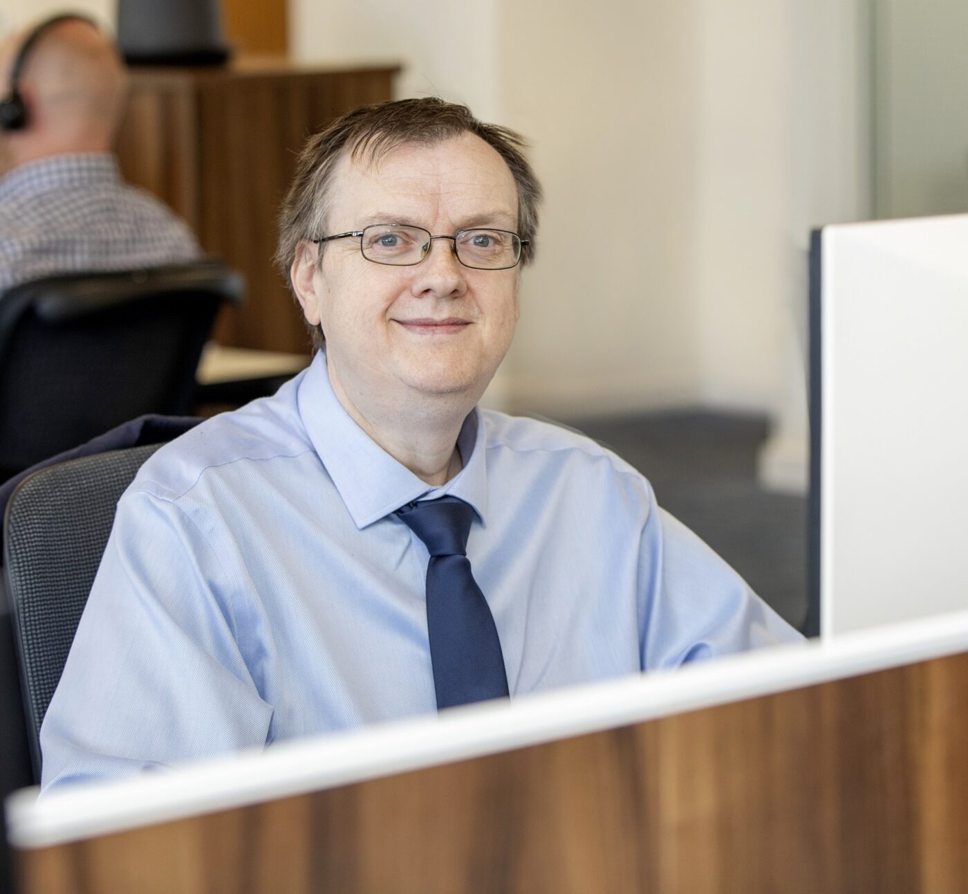 Man in glasses wearing blue shirt and blue tie sitting at his computer smiling at the camera