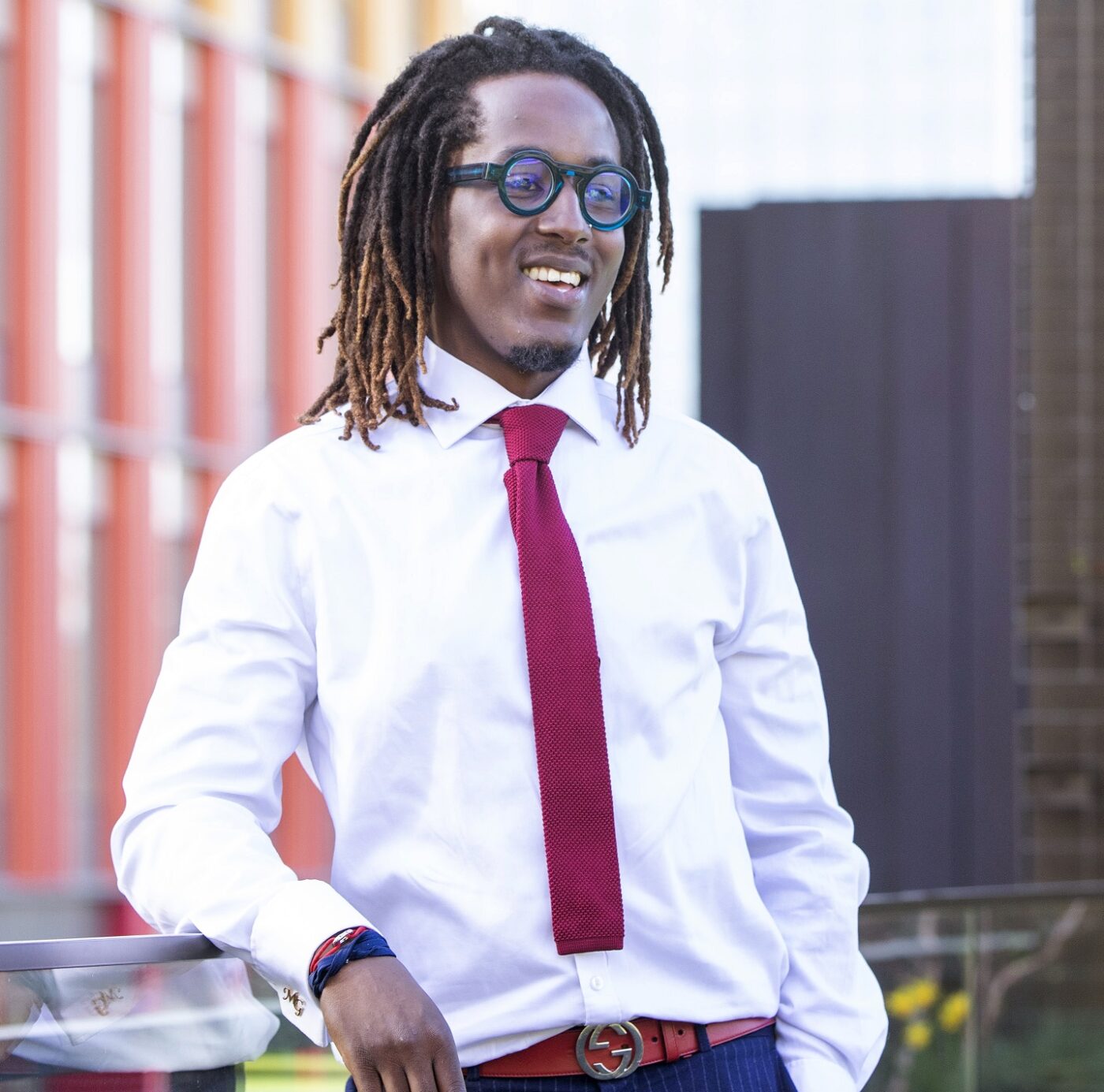 Man in glasses wearing white shirt and red tie leaning on balcony with red office building in background