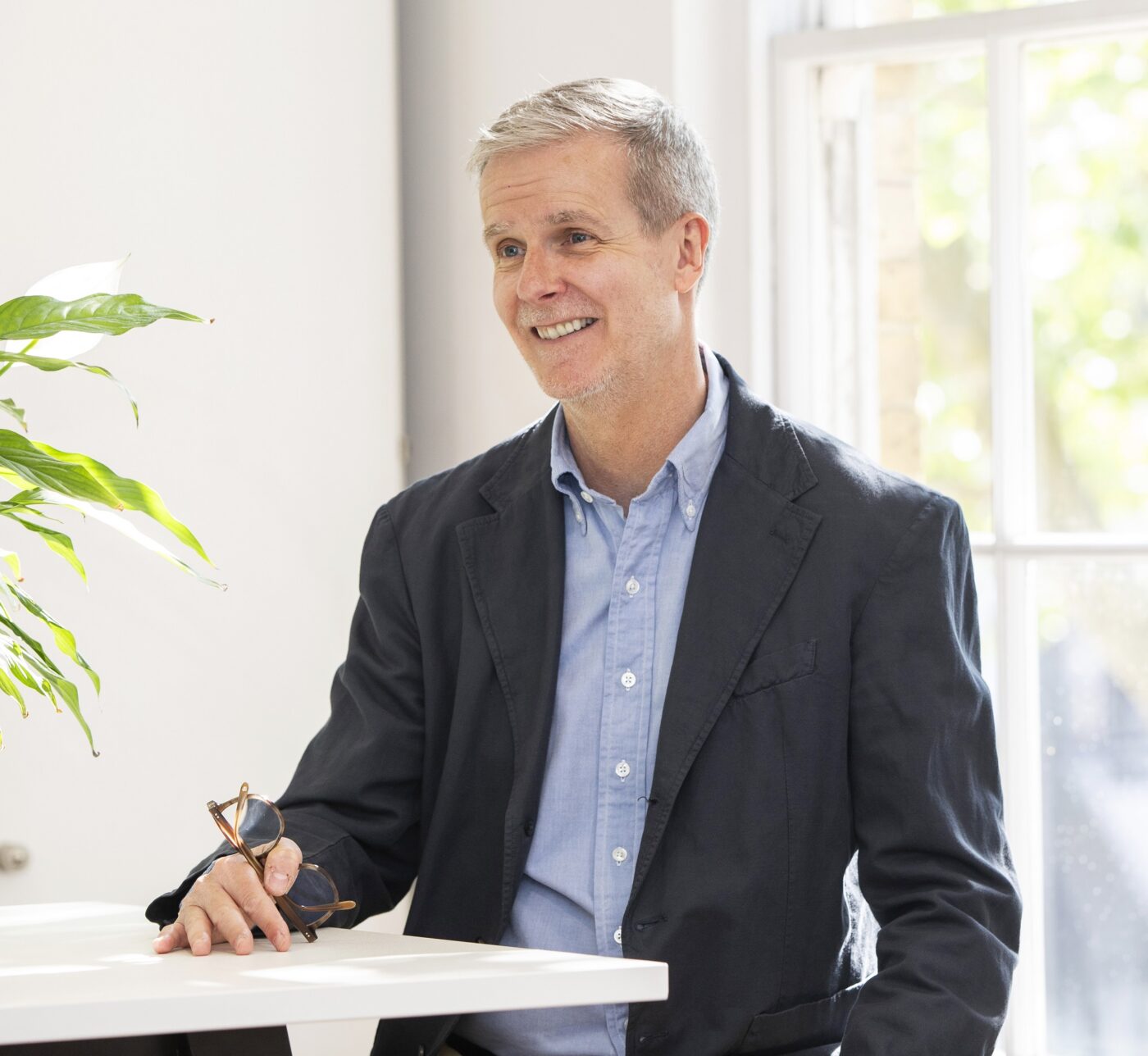 Man in a sunny office space wearing a blue shirt and grey jacket with his glasses in his hand