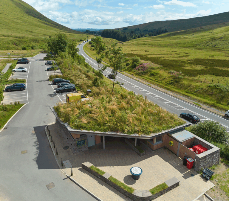 Aerial view of an access point with parked cars, a few buildings, and a stream running through green fields and forest.