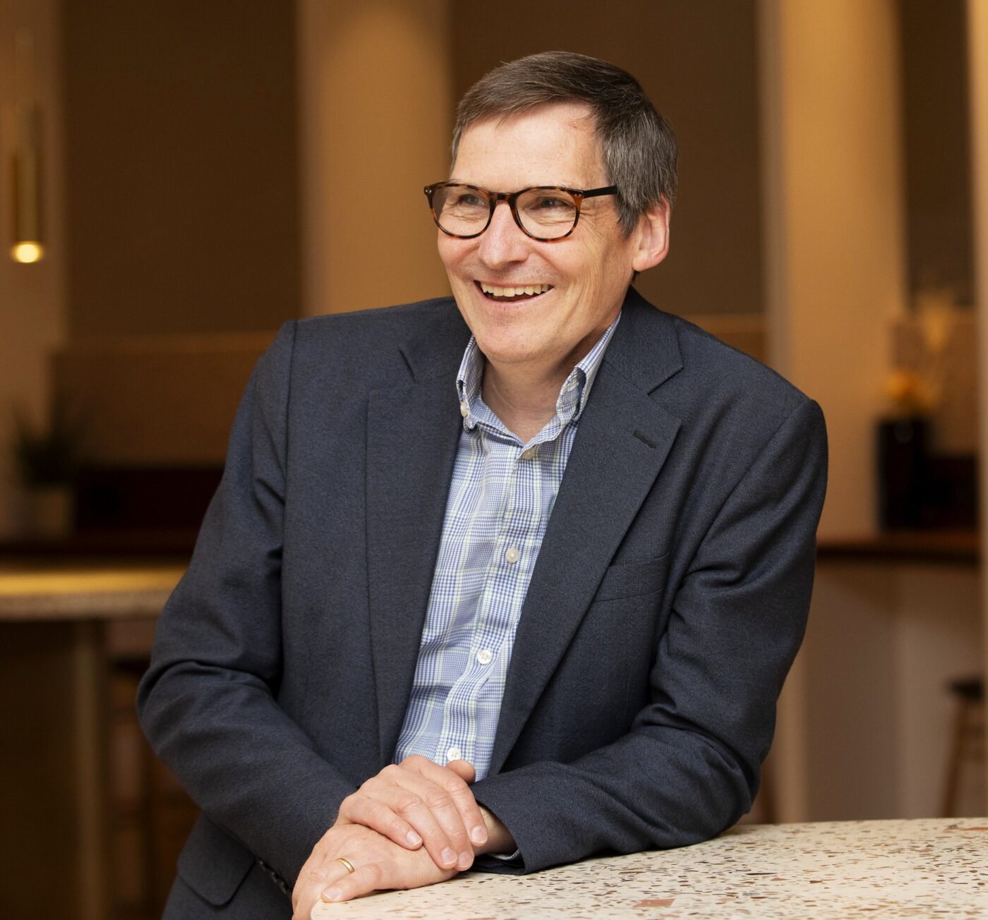 Man with glasses in blue shirt and navy jacket resting on a sideboard