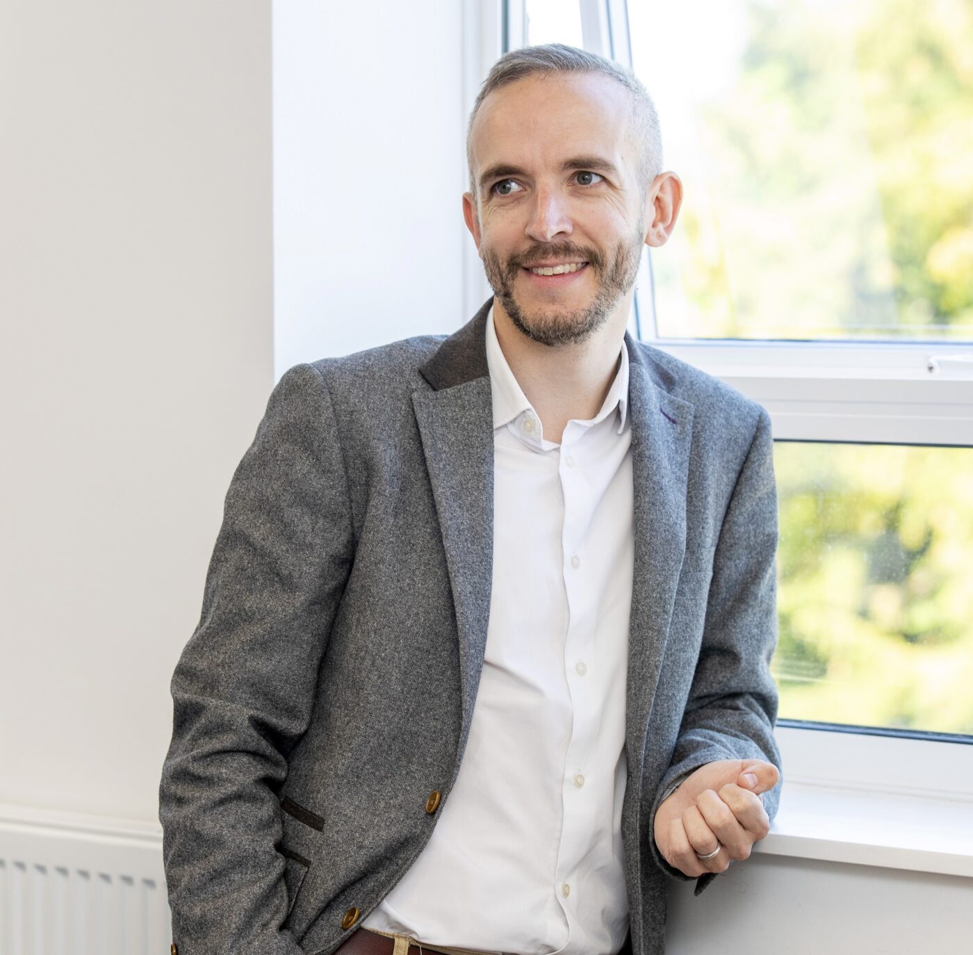 Man in white shirt and grey jacket leaning on a windowsill.