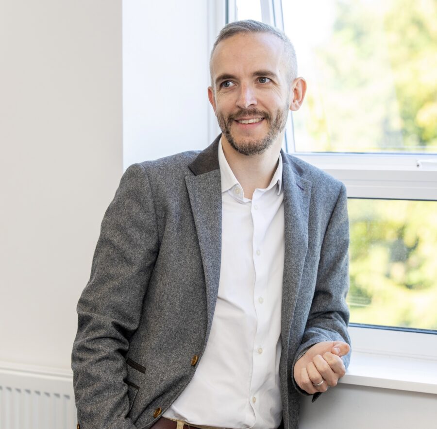 Man in white shirt and grey jacket leaning on a windowsill.