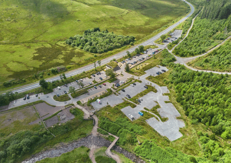 Aerial view of an access point with parked cars, a few buildings, and a stream running through green fields and forest.